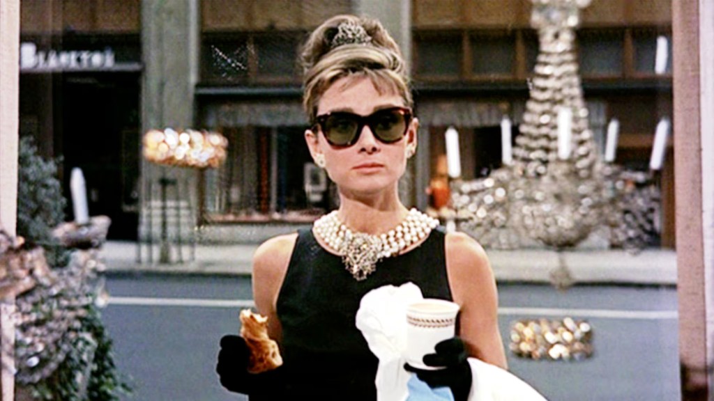 A thin white woman with large sunglasses holds a paper coffee cup. Her hair is upswept, and a thick layer of pearls is around her neck. She is wearing a sleeveless black dress.