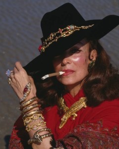 A middle-aged brunette, wearing a hat cocked over her right eye, stares into the camera. A slim cigar in a holder is in her mouth. A great deal of ash is on the edge of the cigar. Her arm, covered in gold bracelets, is out in front of her. A gold necklace shaped like na alligator is around her neck.